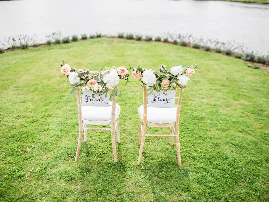 Two white ceremony chairs decorated with floral arrangements on grass.